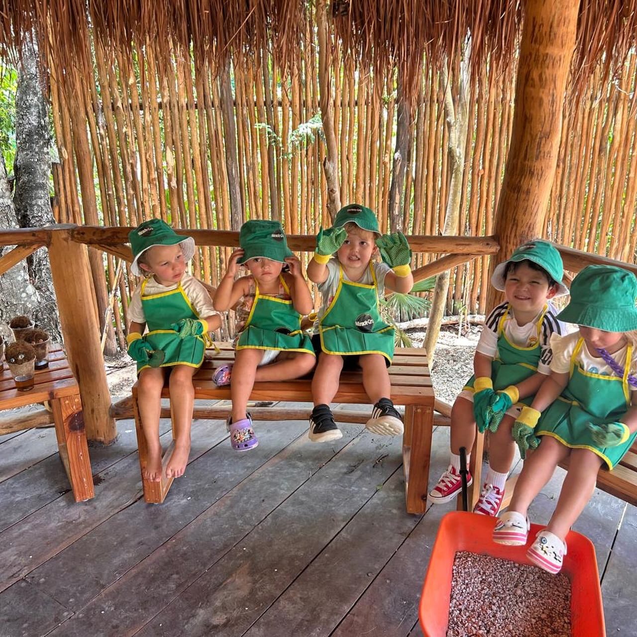 Niños de maternal con sombrero y delantal verde sentados en la palapa